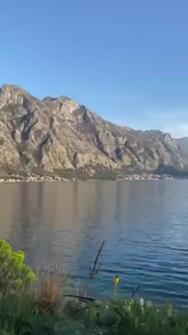 The video shows a view of a bay surrounded by mountains. The camera pans to the left, revealing a road running along the water's edge. A person's shadow is visible on the road, suggesting they are holding the camera and walking. The sky is clear and blue, and the landscape is bathed in sunlight. The mountains are rocky and rugged, with sparse vegetation. The water is calm and reflects the sky.