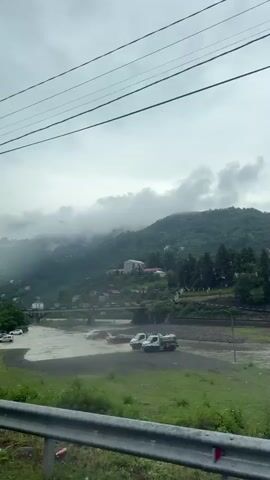 The video shows a view of a river in a hilly, mountainous area. The sky is overcast with clouds. Several cars are visible near and in the river, suggesting potential flooding. A bridge spans the river in the background, and buildings and trees are scattered across the hillsides. Power lines are visible in the foreground and overhead. The camera appears to be moving, possibly from a vehicle, as evidenced by the blurred foreground and the railing that frames the bottom of the shot.