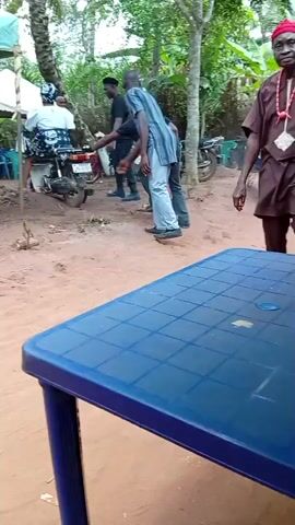 The video captures an outdoor scene in Umundugba, Nigeria, during the daytime. The setting appears to be a casual gathering area with reddish dirt ground, shaded by trees and white canopies. Several individuals are seen moving around.

In the foreground, a blue checkered plastic table occupies much of the frame. A man in traditional brown attire with red beaded necklaces and bracelets walks past the table from right to left. An ambulance siren is audible in the background.

Further back, a dark-colored Toyota Land Cruiser SUV, identified by "V8 4WD" on its rear, is parked on the dirt path. Another man, wearing an orange T-shirt with "GUCCI" emblazoned on it and light brown pants, walks past the SUV, smiling and gesturing towards the right. Other people are visible in the background, including a person sitting on a motorcycle near a white canopy, and others standing or walking further down the path. The video ends with a close-up shot of feet walking on the dirt ground. The atmosphere suggests a relaxed, social outdoor event.