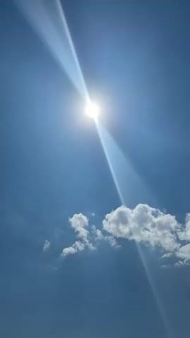 The video shows a clear blue sky with the sun shining brightly and casting a beam of light downwards. Below the sky, there is a body of water with gentle waves. In the water, there are a few people. One person is on a green inflatable object on the left side of the frame, and two other people are standing in the water in the center of the frame. A few small white clouds are visible in the sky. The overall scene depicts a sunny day at the beach or a similar body of water.