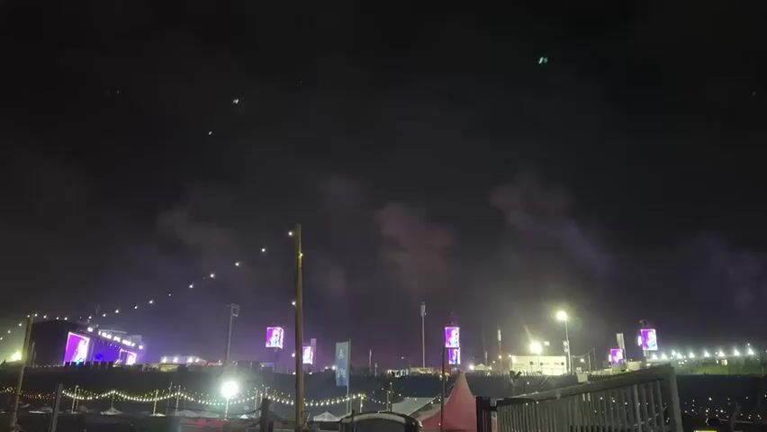 The video shows a night view of an outdoor event. In the foreground, there are strings of small, glowing white lights draped over what appear to be fences or structures. Behind these lights, there's an area with more ground-level lighting and some structures that might be tents or stalls. Further back, there's a large stage to the left, illuminated with bright pink and purple lights. Several tall, rectangular screens are visible, displaying colorful, dynamic visuals. In the background, there's a large area, possibly an open field, with numerous small, green lights scattered across the sky, which could be drones or fireworks. There are also plumes of smoke or fog, illuminated by red and purple lights, rising into the night sky from what appears to be part of the event area. The overall impression is that of a lively night festival or concert.