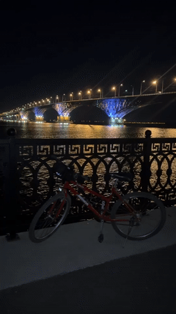 Red bike parked by Volga River near illuminated Saratov Bridge