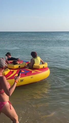 A woman in a pink bikini and a grey baseball cap stands at the edge of the water, holding a black smartphone and looking out at the ocean. She is facing away from the camera. In the water, a yellow and red inflatable tube with "RIF" written on it is floating. Three people are seated on the tube, facing away from the camera. One person on the far left, wearing a blue shirt, has their legs up. The second person, in the middle, has their legs up as well. The third person, on the far right, is wearing a yellow top and has their legs up. The water is calm, and the sky is clear.
