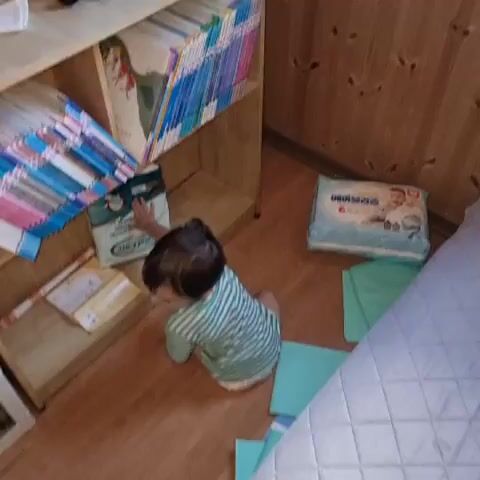 A young child wearing a green and white striped shirt and diaper is sitting on a wooden floor next to a wooden bookshelf. The bookshelf is filled with books. To the right of the child, there is a package of baby wipes and some green cloths. The child turns to look up at the bookshelf.
