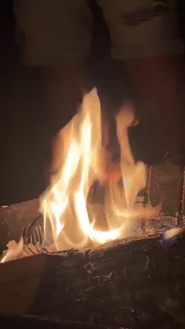 A close-up shot captures bright, flickering flames dancing over burning logs in what appears to be a fire pit or grill. The scene is dark, suggesting nighttime, with the fire providing the main illumination. Some smoke is visible rising from the burning wood. In the background, unseen individuals are talking in Russian. A male voice says, "My daughter acts like we're filming something," and a female voice responds, "Loves it, right?" and then starts to say, "Ruslan, when you're going to...". The overall atmosphere is relaxed and conversational, centered around the warmth and light of the fire. The location is Веревское, Russia.