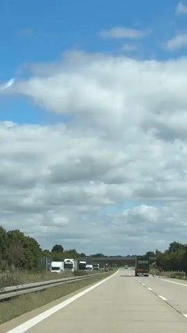 The video shows a view from the passenger seat of a vehicle driving on a highway. The highway has multiple lanes and is bordered by a grassy median and trees. Several trucks are visible on the highway, including a green truck in the right lane ahead and a white truck in the left lane. The sky is blue with large, fluffy white clouds. A bridge crosses over the highway in the distance. The overall scene is of a typical highway drive on a cloudy day.