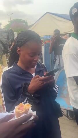 A young woman with braided hair is using a smartphone and holding a pastry in her left hand. She is wearing a navy blue jacket with white stripes on the sleeve. Behind her, a man wearing a black shirt with the word "STORN" printed on it covers his mouth with his hand. A woman wearing a teal hijab walks past on a blue and white metal seat. The scene appears to be outdoors in a crowded area, possibly a market, with tents and other people in the background.