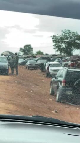 The video shows a view from inside a car driving on a road with cars parked on the shoulder, under a cloudy sky. Rain or water is on the windshield, obscuring the view to some extent. People are visible near the parked cars, some holding umbrellas. The road also has other traffic including motorcycles and what appears to be a tricycle. The environment includes buildings and what seem to be market stalls in the background.