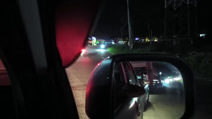 The video shows a view from inside a car at night. The road ahead is visible, with a line of brightly lit vehicles in the distance. A side mirror shows traffic behind the car. There are utility poles and vegetation along the side of the road.