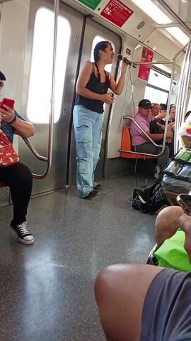 The scene takes place inside a public train, likely a subway, with passengers seated along the sides. A young woman with braided hair, wearing a black tank top and blue cargo pants, stands near a door and sings into a microphone. A man wearing a plaid shirt and ripped blue jeans pushes a heavily laden trolley past her, obstructing the view briefly. The trolley is covered in items for sale, including candy and decorations. Other passengers are visible, some looking at their phones or observing the scene. The train car has grey floors and white walls, and various signs are visible. The overall atmosphere suggests a typical scene of urban life in a location where street vending is common.