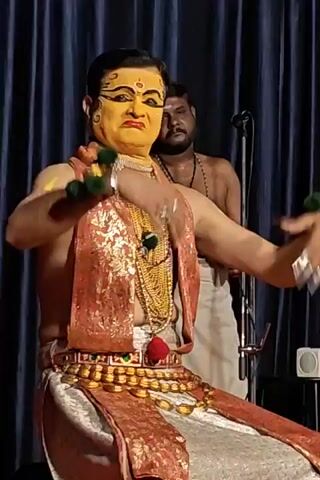 Here's a description of the video:

A male performer in traditional Kathakali costume and makeup sits center stage against a dark blue curtain. The man has yellow face paint with black eyeliner, a gold necklace and belt, and a colorful scarf or shawl draped over his shoulders. He makes gestures with his hands, demonstrating intricate mudras (hand gestures) common in classical Indian dance. He has green and yellow pompoms on his wrists. Behind him, out of focus, stands another man wearing a white garment and a necklace of beads. A microphone stand is also visible in the background. There is a small sound system on the floor in the bottom right. The lighting is bright, illuminating the performer and his costume.