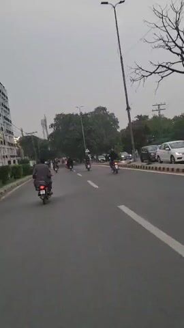 A first-person perspective video captures a street scene with multiple motorcycles driving on an asphalt road under an overcast sky. On the left side of the road, there's a building with white facades and manicured bushes. A white pedestrian walkway is visible along the road. Several trees line the right side of the road, with parked cars visible further down. A few pedestrians are seen on the walkway and crossing the street. The motorcycles, with riders, are proceeding forward on the road, with some appearing to be in motion and others stationary. The overall impression is of a busy street with ongoing traffic.