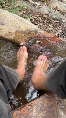 A person is sitting with their feet in a shallow stream of water. The stream is flowing over a flat, rocky surface covered with fallen leaves. The surrounding environment is a forest, with trees visible in the background. The person is wearing dark-colored pants. The water appears clear enough to see the bottom of the stream.
