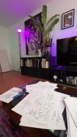 The video shows a person standing in a room with eclectic décor, including artwork, plants, and a keyboard on a stand. The room is lit with colored lights, mainly purple and green. The floor is covered in what appears to be wood paneling. A coffee table in the foreground is covered with papers that seem to be sketches or designs. Pillows bearing the Red Bull Racing logo are visible on a black couch. The person in the room is wearing a black outfit and a baseball cap. The camera moves slightly, changing perspective and showing more of the room.