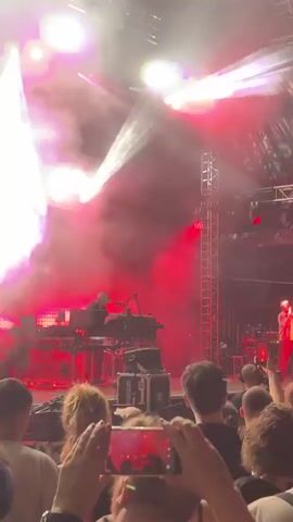 The video shows a dimly lit concert venue. In the foreground, the backs of several audience members are visible, with one person holding up a smartphone to record the event. The stage is set up with various electronic music equipment, including synthesizers, keyboards, and mixers. A person wearing a white hat is operating the equipment. Bright stage lights illuminate the smoke-filled air, creating dramatic beams of light. The overall atmosphere is that of a live electronic music performance.