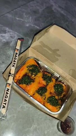 The video shows a top-down view of a food container filled with sushi rolls. The sushi rolls are topped with a vibrant orange sauce and sprinkled with green herbs. A pair of chopsticks with the brand name "NAGATA" printed on them is placed next to the container. To the right of the container, a small plastic cup filled with a dark sauce is visible. The food container is placed inside a brown cardboard box, which is open, revealing a slightly wet interior. The scene is set on a grey, marble-like surface. The lighting is dim, suggesting an indoor setting, possibly at night. The video is a close-up shot of the food, with the camera slowly panning over the sushi.