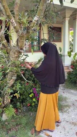 A person wearing a black hijab and a mustard-yellow pleated skirt stands in front of a tree with vines and small pink and red flowers. They are reaching out to touch a purple flower on a bush to the right of the tree. The background shows a portion of a house with a window and pillars. The ground is covered with dirt, leaves, and grass. The lighting suggests it is daytime.