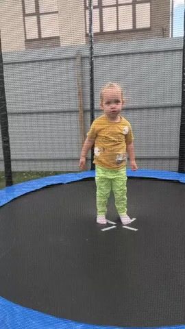 A young child with blonde hair and a yellow t-shirt and green pants is on a trampoline. The child is standing with their feet apart and arms slightly out to the sides. The child then jumps up and down several times. The trampoline has a blue edge and a black jumping surface with white markings. A safety net surrounds the trampoline, and a gray fence is visible in the background. The child appears to be playing.