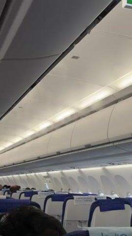 The video shows the interior of an airplane cabin. The camera is positioned looking up towards the ceiling where the overhead compartments are located. A fine mist of water vapor is being released from the air vents along the ceiling, creating a visible fog. Several rows of airplane seats, upholstered in blue and white, are visible below the ceiling, with passengers seated in some of them. The lighting in the cabin appears to be a soft, warm white. The overall impression is a calm and orderly cabin environment during flight.
