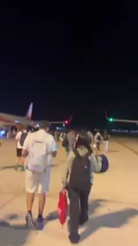 At night, passengers are walking from planes across an airport tarmac. One plane, labeled “jet2holidays,” is parked. Ground staff are present, and airport vehicles can be seen nearby.