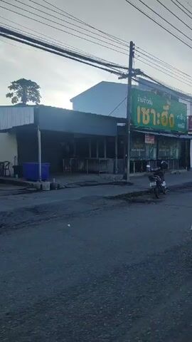 The video shows a street scene in Thailand from a motorcycle's perspective. A woman on a motorcycle drives past a restaurant with a large green sign. The street is lined with buildings and various parked cars and trucks. Several people are walking along the sidewalk and crossing the street. The weather seems overcast. Electrical wires are visible overhead.