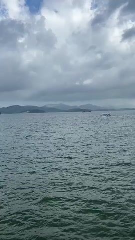 The video shows a body of water with a boat moving from left to right across the frame. The water is choppy, and the sky is cloudy with mountains in the distance. The boat appears to be a speed boat with a few people on board. The overall impression is that of a cloudy day at sea.