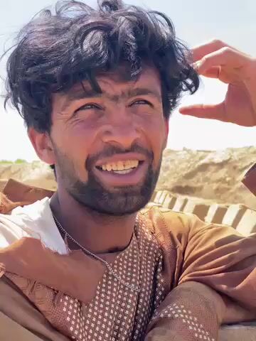 A close-up shot shows a man with dark, curly hair and a beard, smiling and looking slightly off-camera. He is wearing a light-colored top under a brown patterned garment and a silver chain around his neck. Another hand enters the frame briefly to gently touch his hair. The man appears relaxed and engaged in conversation. The background consists of bright, sandy or earthen mounds under a clear, bright sky, suggesting an outdoor, possibly arid or construction-related setting.
