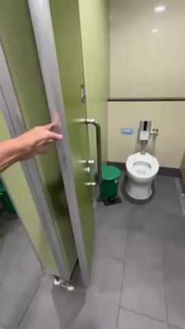 A person's arm and hand are shown in the foreground, pushing open a bathroom stall door with a metal handle and frame. The door is light green, and the surrounding stalls are the same color. Inside the opened stall, a white toilet is visible, along with a green trash can. The walls are beige and gray tiles cover the floor. The camera angle is slightly downward, focusing on the door and the inside of the stall.