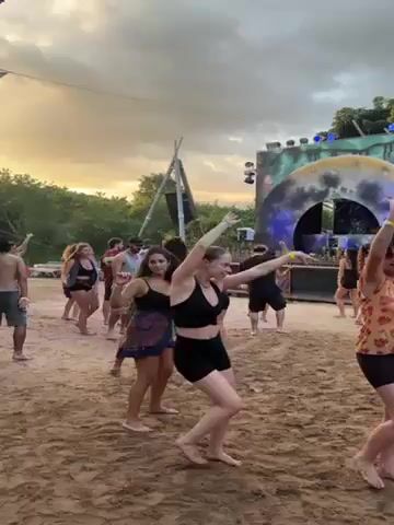 The video shows a group of people dancing to music at an outdoor event. The ground is sandy, and there's a stage with speakers.  A large, colorful, canopy covers much of the area.  There are tents and other structures set up, and people can be seen walking around and talking.  The sky is cloudy, and the sun appears to be setting. The overall atmosphere is festive and energetic.