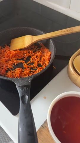 The video shows someone cooking in a kitchen. A speckled black frying pan is on a black glass stovetop. Inside the pan, thin strips of shredded carrots are cooking. A wooden spatula with the word "natura" inscribed on it rests on the edge of the pan. Next to the stovetop, there's a small wooden bowl and a white bowl filled with a red liquid. A hand pours the red liquid from the bowl into the pan, covering the carrots. Steam rises from the pan as the mixture begins to bubble. The hand then uses the wooden spatula to stir the carrots and red liquid together. The kitchen appears clean and well-lit.