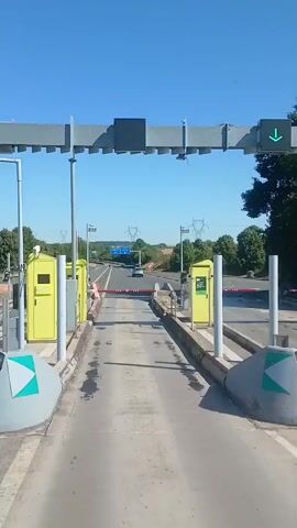 A video shows a view from inside a vehicle approaching a toll booth on a clear, sunny day. The video begins with a green arrow indicating the lane is open. As the vehicle gets closer, a red barrier arm obstructs the lane. The barrier arm is white with red reflective stripes. To the left and right of the lane are yellow toll booths. The road ahead is visible with trees and power lines in the distance. The video ends as the vehicle is about to pass through the toll booth.