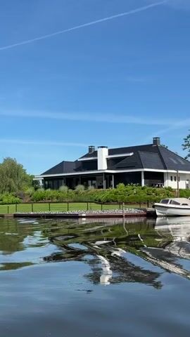 The video shows a shot of a canal in a town. On the left side of the canal are houses and boats. The houses have dark roofs and white walls. There is a green lawn and various shrubs. The water is reflective, showing the outline of the houses and boats on its surface. The camera slowly pans towards the right, and the backdrop changes to tall grass and trees. The sky is a vibrant blue with a few wispy white clouds.