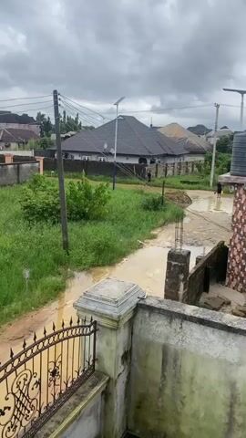 The video shows a street scene with cloudy skies and evidence of recent rain, as puddles of water are visible on the unpaved road. In the foreground, a partially constructed wall with metal fencing is visible. Beyond this, the road curves to the left, bordered by overgrown green grass and bushes. A utility pole with wires stands to the left of the road. In the background, several houses with dark-tiled roofs are situated behind low walls. One house has a solar panel on its roof. Further down the road, two people can be seen walking. To the right of the road, there is another building with a patterned facade and a large black water tank on top. Another person is walking away from the camera on the right side of the road, wading through water. The overall impression is of a rural or semi-urban area experiencing wet weather.
