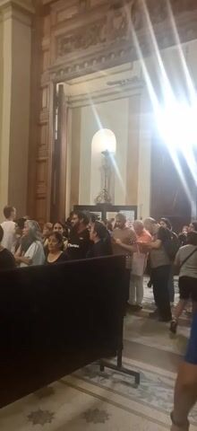 The video shows the interior of a large, ornate church filled with people. The camera pans upward from a crowd of people towards the front of the church, revealing high, arched ceilings, marble pillars, and elaborate decorations. Many people are milling around or sitting in pews, facing the altar. The church is well-lit, with beams of light streaming through the high windows. There are display screens at the side of the church.