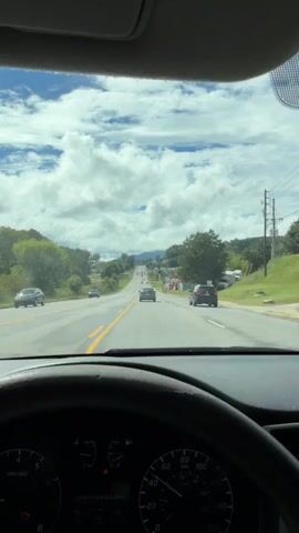 The video is taken from the driver's perspective inside a car. The car is on a paved road with two lanes in each direction, separated by a yellow double line. The road is going uphill and curves to the left. There are other cars on the road, driving in the same direction as the camera car. On the sides of the road, there are trees, bushes, and some buildings, including what appears to be a gas station or convenience store. The sky is blue with large, puffy white clouds. The dashboard of the car is visible in the foreground, showing various gauges and indicators. The steering wheel is also partially visible. The overall impression is of a scenic drive on a sunny day.