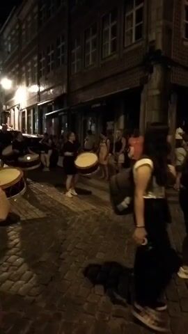 A group of people are playing drums in the street at night. The street is paved with cobblestones and lined with buildings. Some people are standing and watching, while others are playing the drums. The lighting is dim, creating shadows on the street. The people playing the drums are wearing dark clothing, and the drums are large and round with light-colored drumheads. One person in the foreground is wearing a dark t-shirt and shorts and is actively playing a drum. The person to their left is also playing a drum. Behind them, a few more people are playing drums. A few spectators are visible in the background, some standing and some sitting.