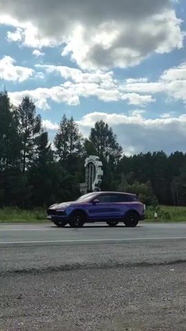 The video shows a roadside sign in a natural setting. The sign is tall and appears to be made of metal, with a large gear-like structure at the top. Inside the gear, there is a latticework structure. The sign has Russian text inscribed on it, which appears to say "ДОБРОГО ПУТИ" (Dobrogo Puti), meaning "Have a good trip". The sign is located next to a paved road. In the background, there is a dense forest with various trees, and the sky above is partly cloudy. A small red and white pole is visible in the distance to the right of the sign. On the left side of the video, a portion of a red and white billboard is visible. A red vehicle with a trailer drives past the sign on the road, moving from right to left. The video is shot from a moving vehicle.