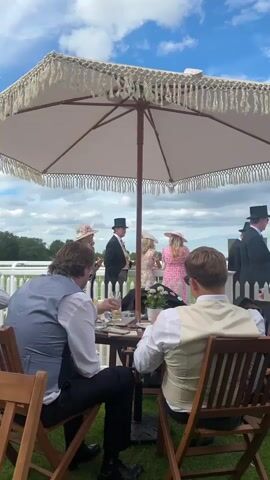 The video shows a group of people dressed in formal attire at an outdoor event, possibly a horse race. In the foreground, two men are seated at a small table under a white fringed umbrella, facing away from the camera. Both men are wearing waistcoats over white shirts, with one man in a gray waistcoat and the other in a tan waistcoat. The man in the gray waistcoat is wearing dark pants and the man in the tan waistcoat is wearing lighter pants. Their backs are to the camera as they look towards a group of people in the background. The table has drinks and what appears to be food on it. In the background, several people are standing near a white fence. One man is wearing a top hat and a suit, and two women are wearing hats and dresses. The sky is blue with some clouds. The ground is covered in grass.