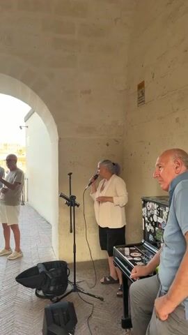 A woman with gray hair sings into a microphone while standing under an archway. A man in a light-colored shirt and white shorts stands in the background holding a phone. Several other people are visible in the background near a railing, looking out at a cityscape. A keyboard and speaker are on the right side of the frame.