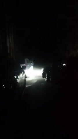 The video shows three cars parked on what appears to be a narrow road at night. The headlights of the middle car are on, illuminating the road. The other two cars are silhouetted in the darkness, making it difficult to see details. The background is a dark, wooded area. Someone can be heard speaking in the distance. The overall scene is dark and somewhat mysterious.
