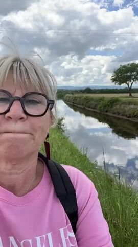 A woman with short gray hair and glasses stands next to a narrow canal on a cloudy day. She is wearing a pink sweatshirt with the word "ANGELES" printed on it, and has a black backpack. The canal is surrounded by green grass and weeds, and reflects the sky. In the distance, there is a field with a lone tree, and mountains.