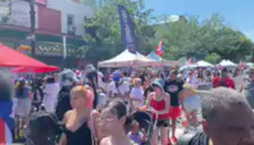 This media file captures a lively outdoor event, likely a festival or street fair, on a bright, sunny day. The scene is bustling with a large crowd of people of diverse ages and ethnicities mingling, suggesting a celebratory atmosphere. 

Dominating the foreground and midground are numerous individuals, many of whom appear to be enjoying themselves. Some are dressed in casual summer attire, while others wear festive clothing, including headwear and vibrant colors. The presence of multiple tents suggests vendors or stalls, possibly selling food, crafts, or other goods. A prominent Puerto Rican flag is visible, indicating a potential cultural celebration or gathering.

The setting appears to be an urban environment, with buildings and trees forming the backdrop. The bright sunlight and clear sky imply fair weather, contributing to the outdoor nature of the event. The overall mood seems to be one of community engagement and enjoyment, with people interacting and moving through the space.

While no specific text is clearly legible on signs or clothing, the visible Puerto Rican flag is a significant cultural identifier for the event. The image conveys a sense of vibrancy and festivity, characteristic of a public celebration or cultural gathering in New York City.