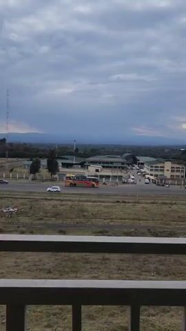 The video shows a slightly elevated view of a multi-lane road in an open area with buildings, vehicles, and distant mountains. There is a rail line in the middle ground, dividing a grassy field from the road. The road features various vehicles, including a long multi-colored bus, cars, and trucks. In the background, several buildings can be seen, interspersed with trees. On the horizon are mountains under a sky with a cloudy overcast. A portion of a railing is visible in the foreground, indicating the view is from an elevated structure like a balcony or rooftop. A person is walking on the grassy field.