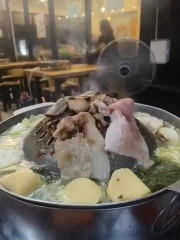 The media file captures a close-up, low-angle shot of a sizzling hot pot filled with various ingredients. The primary focus is on the central dome-shaped grill, where pieces of meat, likely pork, are being cooked. Surrounding the grill, the broth is bubbling vigorously, with ingredients like cabbage, and what appear to be slices of potato or taro submerged within it. The steam rising from the pot indicates the meal is actively being prepared.

In the background, the scene suggests an indoor restaurant setting, possibly an outdoor seating area covered by a roof. Wooden tables and stools are visible, arranged in a casual dining layout. A black electric fan is positioned behind the hot pot, likely to help dissipate the smoke and steam. Silhouettes of people can be seen in the distance, suggesting other diners are present, contributing to the ambient atmosphere of a bustling eatery. The lighting appears to be artificial, suggesting it is evening or night. The overall mood conveyed is one of warmth and hearty dining. No visible text is present in the media file. The location is identified as Chiang Rai City Municipality, Thailand, consistent with the visual cues of Asian cuisine and dining style.