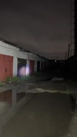 The video captures a nighttime scene in an outdoor alleyway lined with garages. The ground is wet and shows puddles of water. On the left side of the alley, there are several garage doors, some with red fronts. On the right side, there are also garage doors, and further down the alley, a tall metallic tower with a ladder is visible. The sky is dark and overcast, suggesting it might be raining or has recently rained. A faint light beam, possibly from a flashlight, is directed down the alley, illuminating a portion of the ground and the garages. The scene appears deserted.