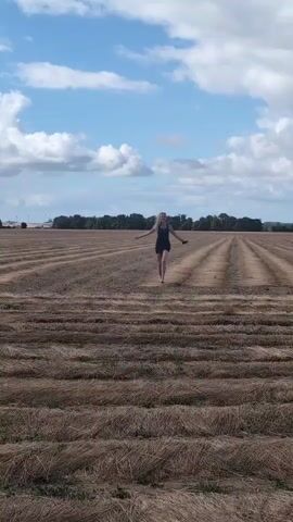 A woman with blonde hair wearing a black dress is walking in a harvested field. The field is covered in rows of dried grass or straw. The sky is blue with white clouds. The woman is facing the camera with her arms outstretched. The video is shaky and appears to be a vertical recording.
