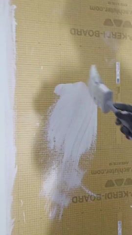 The video shows a close-up of a person's gloved hand applying a white substance to a grid-patterned surface with a brush. The surface appears to be a building material, possibly a backer board for tiling, as indicated by the visible text "KERDI-BOARD" and "SCHLUTER.COM". The person is wearing a dark gray glove and holding a brush with a blue and white handle, applying the white material in broad strokes. The action suggests a construction or renovation project, likely involving waterproofing or preparing a surface for finishing. The lighting is consistent, suggesting an indoor environment. No other people, specific locations, or weather conditions are visible or implied. The text on the board is the most prominent detail besides the action itself, suggesting a brand or product name related to construction.