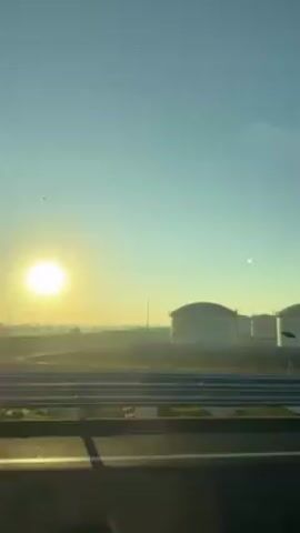 The video shows an industrial area under a clear blue sky with the sun rising on the horizon. Several large, domed storage tanks are visible in the distance, silhouetted against the bright sunlight. The ground in front of the tanks is flat and grassy, with a few small, rectangular structures placed alongside the road. The foreground shows a paved surface with painted white lines, indicating a road or parking area. The view is captured from a moving vehicle, likely a car or bus, suggested by the slight blur and motion. Music is playing in the background.