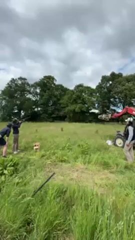 Here's a description of the video:

The video captures a skeet shooting session in a grassy field under a cloudy sky. Three people are positioned to the left of the frame. Two of them are actively engaged in shooting at clay targets launched by a red and gray crane machine that is visible in the distance to the right of the frame. One person observes. They are dressed in casual attire, including jackets and khaki pants. Shotguns are used. Trees surround the field. Tall grass occupies the foreground. A low fence runs across the middle of the grass. A small wooden stand is set up in front of the shooters. A person wearing a white shirt and khaki pants stands by the crane. The sounds of the shotgun blasts and the breaking clay targets can be heard.