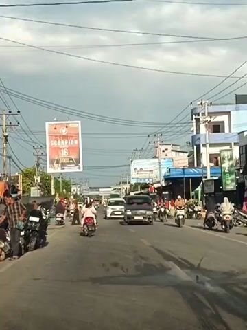 A two-lane asphalt road with moderate traffic. On the left, parked motorcycles and several people are visible. On the right, buildings line the street, with more motorcycles and some people moving about. Overhead, a complex network of electrical wires crisscrosses the sky, which is partially cloudy. A large billboard with red and white text reading "SCORPION 16" is prominently displayed on the left side of the road. Several vehicles, including cars and many motorcycles, are actively moving in both directions on the road. One white car in the right lane has a license plate visible. The overall atmosphere appears to be a typical daytime scene in a populated area.